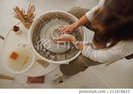 Female potter in apron making shape of clay vase on spinning pottery tool in ceramic workshop 111678633