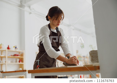 Professional female potter in apron kneads piece of clay with her hands on table in studio 111678763