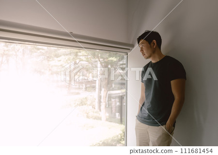 Asian man leaning against a wall, gazes out the window 111681454