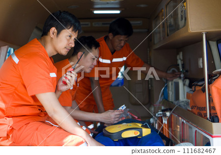 Two male nurse in an orange uniform measure vital signs of an  accident victim on a bed in an ambulance. The other man use walkie talkie to coordinate the nearest hospital. 111682467