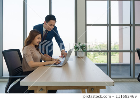 Young asian woman in casual wear typing on laptop computer keyboard. Her boss in suit and tie, stand pointing at the screen, discussing work or collaborating on a project. Young asian woman in casual wear typing on laptop computer keyboard. Her boss in suit and tie, stand pointing at the screen, discussing work or collaborating on a project. 111682469