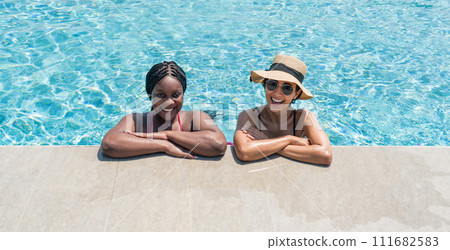 Two women are relaxing together on the edge of a swimming pool, enjoying their time and chatting. Two women are relaxing together on the edge of a swimming pool, enjoying their time and chatting. 111682583