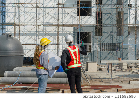 Two construction workers are standing together in front of a large building site with helmet and safety gear. Day time work safety checks. Work environment at the site of housing projects. Two construction workers are standing together in front of a large building site with helmet and safety gear. Day time work safety checks. Work environment at the site of housing projects. 111682696