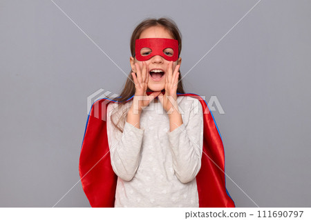 Overjoyed funny positive little girl wearing superhero costume and mask isolated over gray background holding hands near mouth screaming loud making announcement Overjoyed funny positive little girl wearing superhero costume and mask isolated over gray background holding hands near mouth screaming loud making announcement 111690797