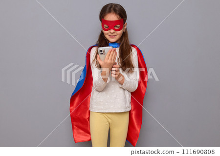 Smiling little girl wearing superhero costume and mask isolated over gray background using mobile phone for scrolling online checking social media browsing web pages 111690808