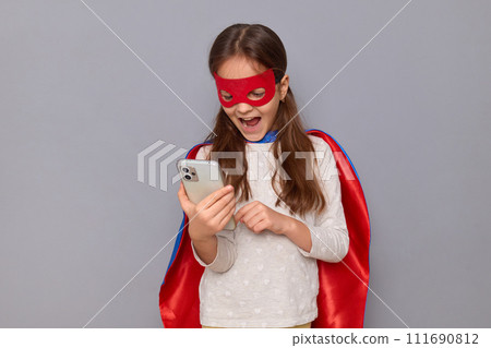 Extremely happy overjoyed excited little girl wearing superhero costume and mask isolated over gray background looking at smartphone screen with open mouth scrolling online Extremely happy overjoyed excited little girl wearing superhero costume and mask isolated over gray background looking at smartphone screen with open mouth scrolling online 111690812