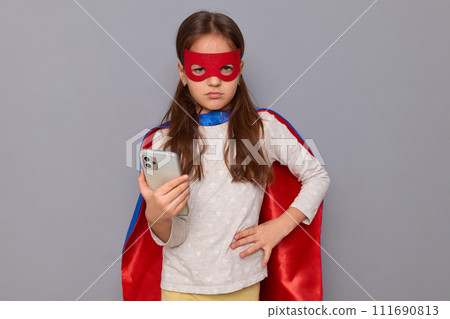 Sad serious little girl wearing superhero costume and mask isolated over gray background keeps hand on hip looking at camera with unhappy face holding smartphone Sad serious little girl wearing superhero costume and mask isolated over gray background keeps hand on hip looking at camera with unhappy face holding smartphone 111690813