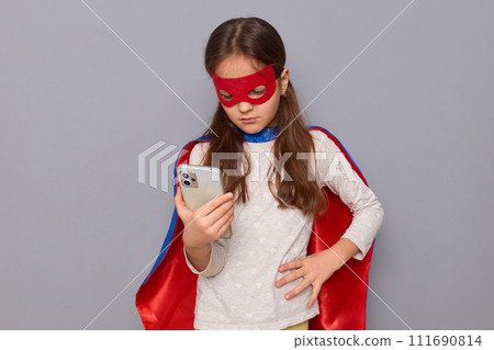 Displeased little girl wearing superhero costume and mask isolated over gray background using mobile phone for online communication looking at screen with upset expression 111690814
