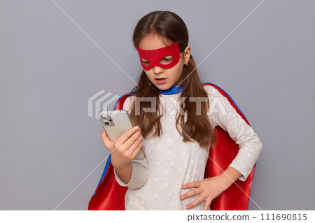 Concentrated little girl wearing superhero costume and mask isolated over gray background reading text message on smartphone holding hand on hip having displeased facial expression 111690815