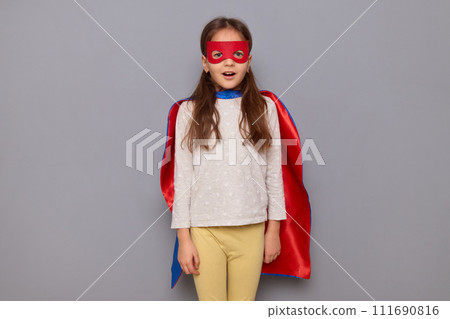 Amazed little girl wearing superhero costume and mask isolated over gray background looking at camera saying wow or omg sees something astonishing being surprised Amazed little girl wearing superhero costume and mask isolated over gray background looking at camera saying wow or omg sees something astonishing being surprised 111690816