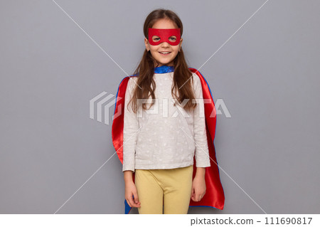 Smiling beautiful little girl wearing superhero costume and mask isolated over gray background looking at camera with happy face expressing positive emotions 111690817
