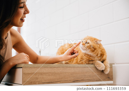 A happy young woman at home embraces her lovely Scottish Fold cat on a table, illustrating the concept of pet love and togetherness. Their affectionate bond shines in this heartwarming portrait. 111693359