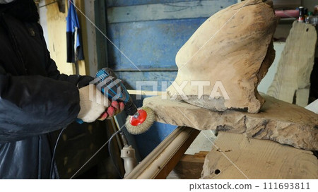The carpenter in gloves holds a drill with a round nozzle with a metal brush to clean off irregularities and roughness on pieces of whole wood in the workshop The carpenter in gloves holds a drill with a round nozzle with a metal brush to clean off irregularities and roughness on pieces of whole wood in the workshop 111693811