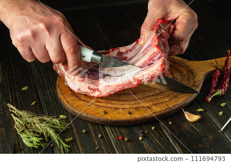 The chef cuts raw ribs with a knife on a wooden board. The concept of the process of preparing a meat dish. Asian cuisine The chef cuts raw ribs with a knife on a wooden board. The concept of the process of preparing a meat dish. Asian cuisine 111694793
