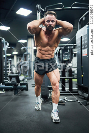 Focused bearded bodybuilder with strong abs, pulling weight, training in gym. Low angle view of handsome determined guy with hands behind head, exercising, working out indoor. Concept of bodybuilding. 111696042