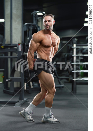 Middle-aged handsome Caucasian male bodybuilder performing side triceps pose in gym. Full length of shirtless athlete showing lateral triceps muscle, looking away. Bodybuilding concept. Middle-aged handsome Caucasian male bodybuilder performing side triceps pose in gym. Full length of shirtless athlete showing lateral triceps muscle, looking away. Bodybuilding concept. 111696045
