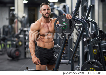 Handsome sportsman posing, while leaning on training apparatus in gym. Front view of shirtless healthy man looking at camera during workout, on blurred background. Concept of sport, lifestyle, health. Handsome sportsman posing, while leaning on training apparatus in gym. Front view of shirtless healthy man looking at camera during workout, on blurred background. Concept of sport, lifestyle, health. 111696046
