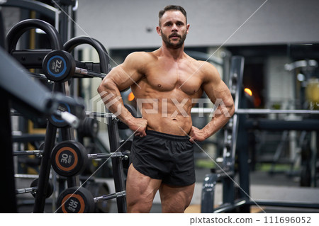 Confident bearded bodybuilder posing with hands on waist in gym. Portrait of strong Caucasian shirtless male athlete, lifting chest up, spreading lats and showing to camera. Concept of bodybuilding. Confident bearded bodybuilder posing with hands on waist in gym. Portrait of strong Caucasian shirtless male athlete, lifting chest up, spreading lats and showing to camera. Concept of bodybuilding. 111696052