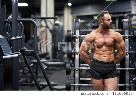 Pensive shirtless crossfit athlete leaning on palf rack barbell platform, having rest after training. Front view of strong, muscular guy looking away, while exercising in gym. Concept of sport. 111696055
