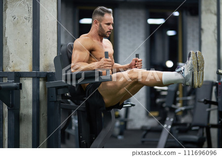 Handsome sportsman with hairstyle pulling up legs, working out indoors. Full body of muscular male athlete in shorts doing pull ups on training apparatus in gym. Concept of sport, workout. Handsome sportsman with hairstyle pulling up legs, working out indoors. Full body of muscular male athlete in shorts doing pull ups on training apparatus in gym. Concept of sport, workout. 111696096
