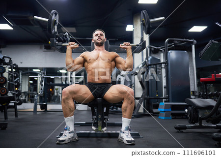 Muscular man with dark hair doing exercise with training apparatus in gym. Low angle view of strong male athlete sitting, using sport equipment during workout. Sport, weight lifting concept. 111696101