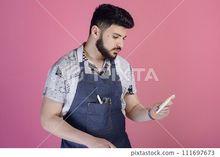 Side view of male designer, hairdresser, barber wearing uniform, holding pen, looking down, thinking. Isolated on pink studio background. 111697673