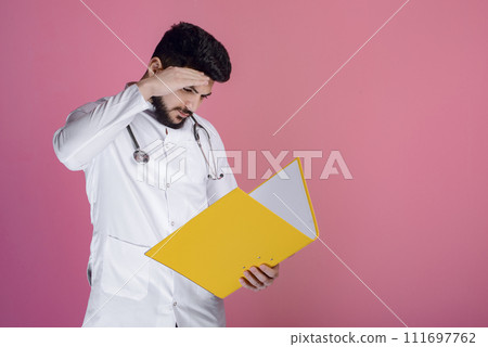 Side view of young doctor with beard standing with stethoscope, holding folder, looking down. Isolated on pink studio background. 111697762