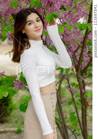 Side view of pretty girl having photoshoot in blossom, holding lilac branch by hands, looking at camera. Concept of springtime. 111697841