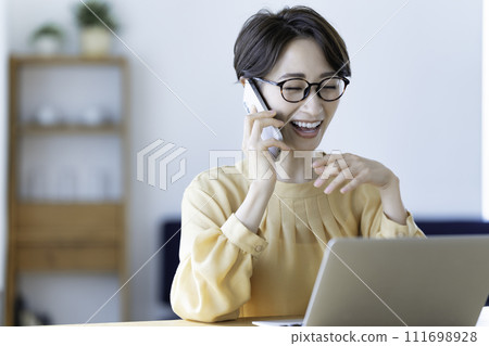 Middle-aged woman talking on a smartphone while using a laptop in the living room 111698928