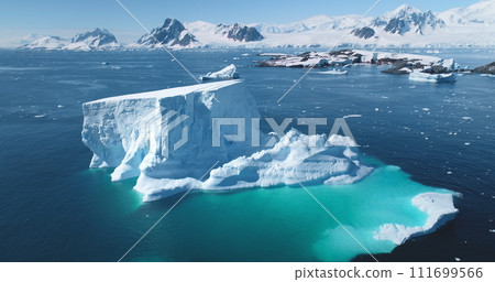 Melting iceberg in Antarctica in sunny day. Huge glacier in turquoise sea water, snow covered mountains in background. Aerial drone flight panorama. Global warming, climate change. Explore South Pole 111699566