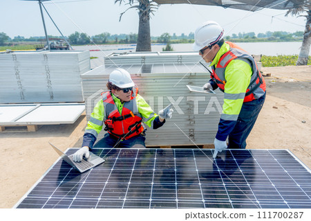 Front view of two Asian professional technician workers one stand and the other sit near solar cell panel and use laptop to discuss about work. 111700287