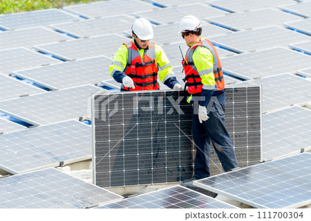 Two Asian professional technician workers with safety uniform stand and help to check or maintenance solar cell panels in area of workplace over water reservoir. Two Asian professional technician workers with safety uniform stand and help to check or maintenance solar cell panels in area of workplace over water reservoir. 111700304