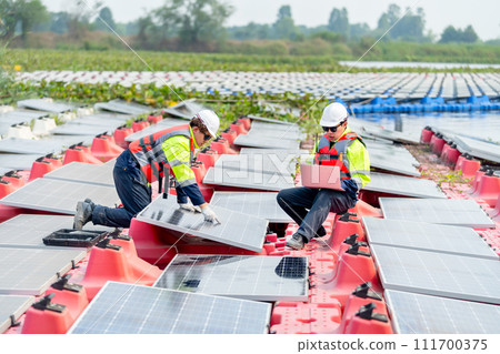 One professional technician worker use laptop to work with other co-worker touch to check and maintenance solar cell panel in workplace over water reservoir. One professional technician worker use laptop to work with other co-worker touch to check and maintenance solar cell panel in workplace over water reservoir. 111700375