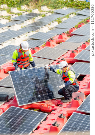 Vertical image of two technician workers help to set up or install solar cell panel into base over water reservoir. 111700386