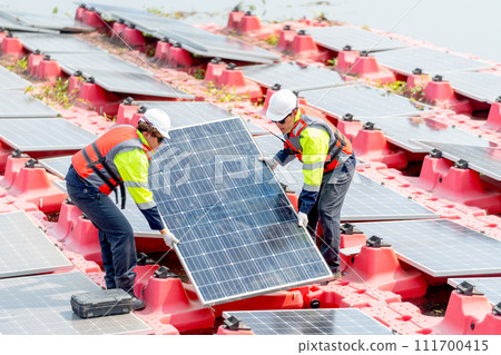 Two professional technician workers hold and carry solar cell panel to set up or install to the base over area of water reservoir. 111700415