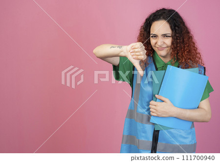 Front view of frown, young woman wearing vest, looking at camera, showing dislike, holding folders. Isolated on pink studio background. Front view of frown, young woman wearing vest, looking at camera, showing dislike, holding folders. Isolated on pink studio background. 111700940