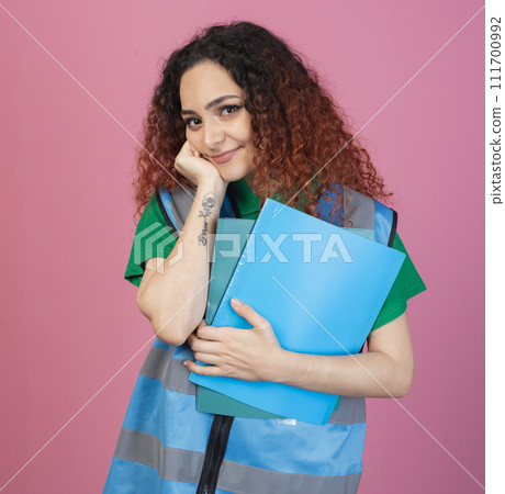 Front view of attractive, young female with red, curly hair standing, leaning, holding folders. Isolated on pink studio background. 111700992
