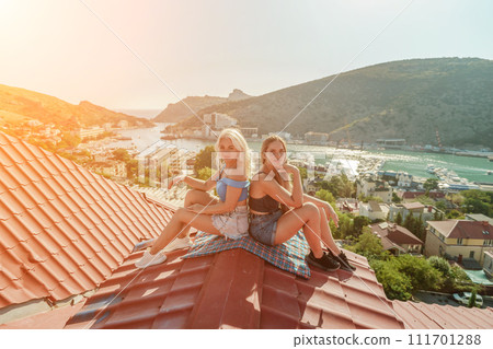 Two women sitting on a red roof, enjoying the view of the town and the sea. Rooftop vantage point. In the background, there are several boats visible on the water, adding to the picturesque scene. Two women sitting on a red roof, enjoying the view of the town and the sea. Rooftop vantage point. In the background, there are several boats visible on the water, adding to the picturesque scene. 111701288