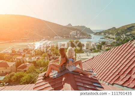 Two women sitting on a red roof, enjoying the view of the town and the sea. Rooftop vantage point. In the background, there are several boats visible on the water, adding to the picturesque scene. 111701435