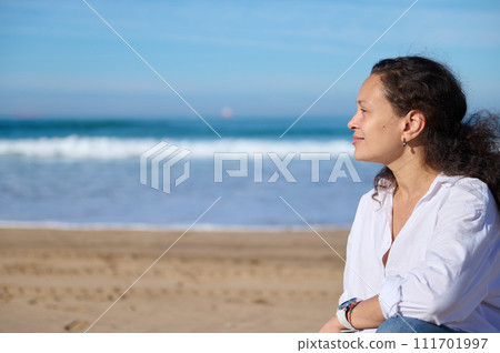 Young woman looking into the distance, sitting alone on the sandy beach against beautiful waves crushing on the sand 111701997
