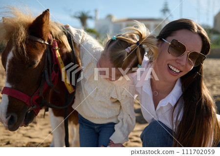 Happy young mother and daughter admiring together a pony horse. Family engaging in outdoors activities with little child Happy young mother and daughter admiring together a pony horse. Family engaging in outdoors activities with little child 111702059