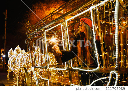 Woman holding sparkler night while celebrating Christmas outside. Dressed in a fur coat and a red headband. Blurred christmas decorations in the background. Selective focus Woman holding sparkler night while celebrating Christmas outside. Dressed in a fur coat and a red headband. Blurred christmas decorations in the background. Selective focus 111702089