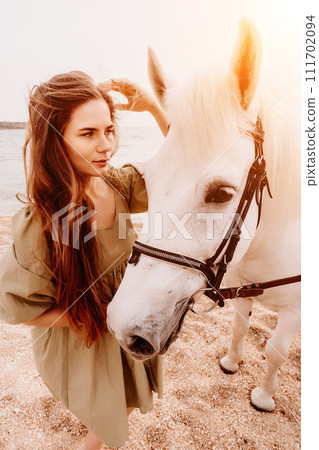 A woman in a dress stands next to a white horse on a beach, with the blue sky and sea in the background. A woman in a dress stands next to a white horse on a beach, with the blue sky and sea in the background. 111702094