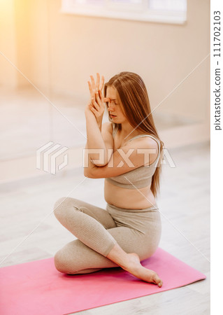 Young woman doing yoga in the gym. A girl with long hair and in a beige tracksuit stands in a cow pose on a pink carpet. A woman performs Gomukhasana. Young woman doing yoga in the gym. A girl with long hair and in a beige tracksuit stands in a cow pose on a pink carpet. A woman performs Gomukhasana. 111702103