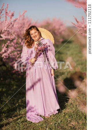 Woman blooming peach orchard. Against the backdrop of a picturesque peach orchard, a woman in a long pink dress and hat enjoys a peaceful walk in the park, surrounded by the beauty of nature. Woman blooming peach orchard. Against the backdrop of a picturesque peach orchard, a woman in a long pink dress and hat enjoys a peaceful walk in the park, surrounded by the beauty of nature. 111702118