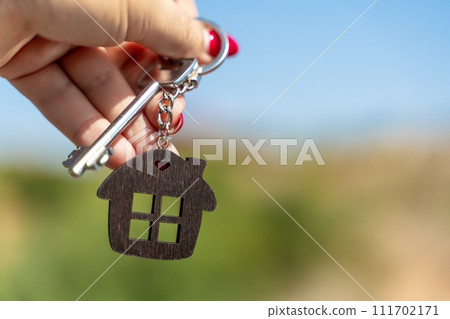Keys with a keychain in the form of a house in a woman's hand against the background of greenery and the sky. The concept of buying a house, apartment, real estate, young family, mortgage loan, rental 111702171