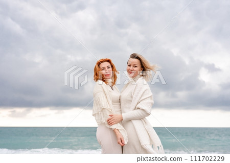 Women sea walk friendship spring. Two girlfriends, redhead and blonde, middle-aged walk along the sandy beach of the sea, dressed in white clothes. Against the backdrop of a cloudy sky and the winter Women sea walk friendship spring. Two girlfriends, redhead and blonde, middle-aged walk along the sandy beach of the sea, dressed in white clothes. Against the backdrop of a cloudy sky and the winter 111702229
