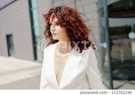 Portrait of a woman standing near a supermarket building. Caucasian model with long dark hair, wearing a white jacket and colored trousers. 111702246