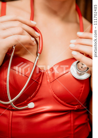 Young woman in red corset and black leather pants with a phonendoscope. Sexy brunette nurse in red and black costume with stethoscope in her hands. Role doctor play. Celebration and party. Having fun Young woman in red corset and black leather pants with a phonendoscope. Sexy brunette nurse in red and black costume with stethoscope in her hands. Role doctor play. Celebration and party. Having fun 111702485