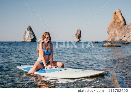 Woman sea sup. Close up portrait of happy young caucasian woman with long hair looking at camera and smiling. Cute woman portrait in a blue bikini posing on sup board in the sea Woman sea sup. Close up portrait of happy young caucasian woman with long hair looking at camera and smiling. Cute woman portrait in a blue bikini posing on sup board in the sea 111702490
